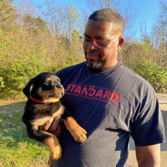 A man outdoors holding a small black and tan puppy with a red collar