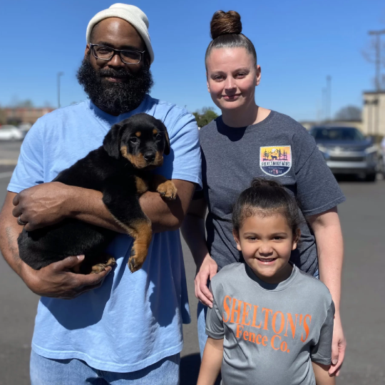 A family standing in a parking lot, one holding a black puppy