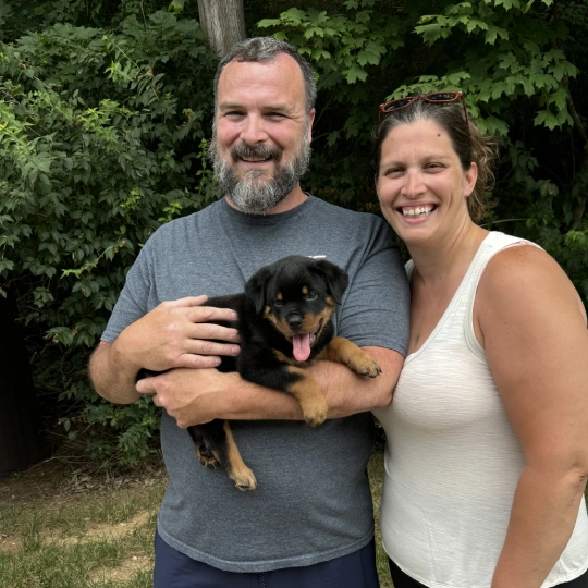 A man and woman standing outside holding a black rottweiler puppy