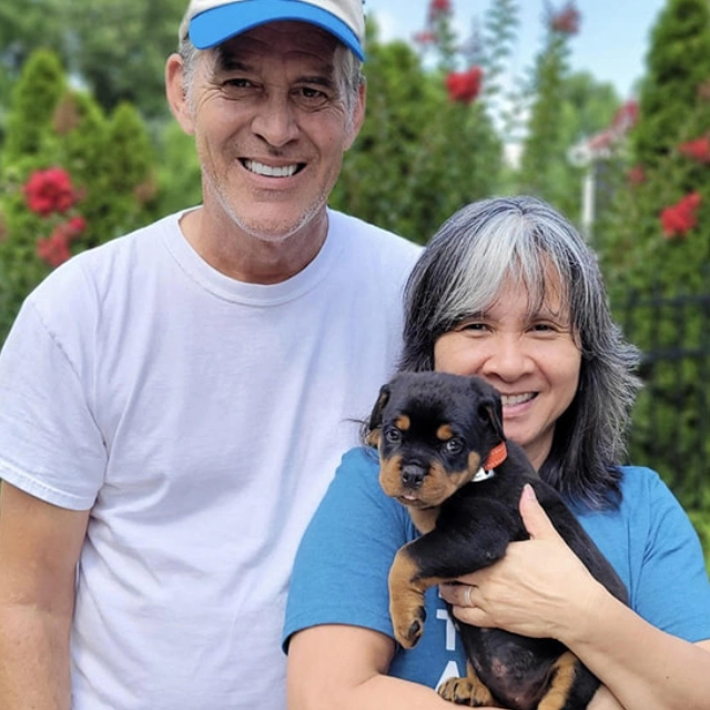 A smiling couple poses outdoors, with the woman holding a small Rottweiler puppy