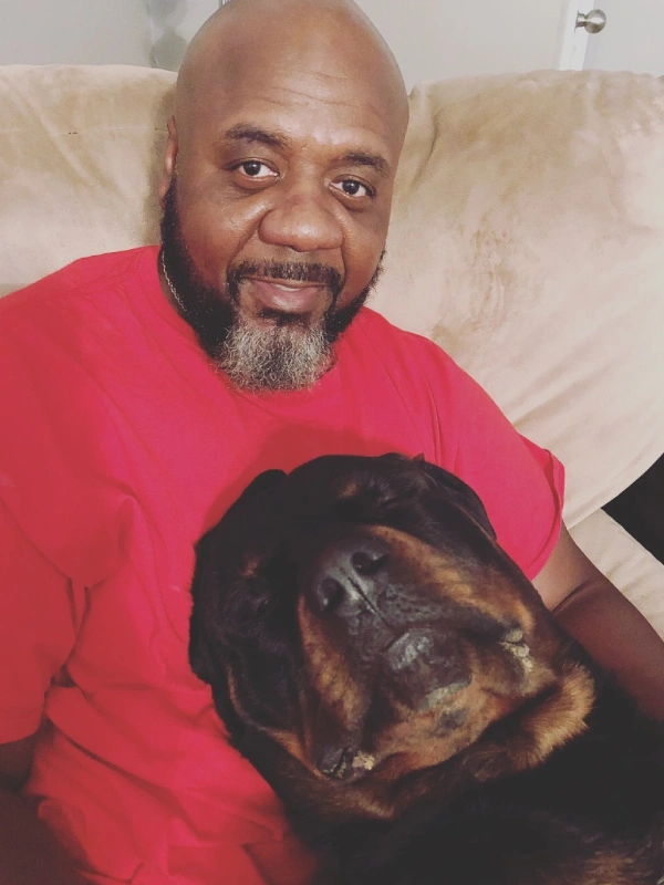 A man in a red shirt sits on a couch with a Rottweiler resting its head on his chest